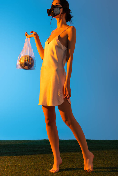 barefoot woman in gas mask holding plastic bag with globe on blue, ecology concept  - Photo, Image