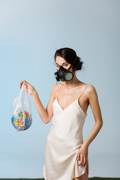 vrouw in gasmasker houden plastic zak met bol op grijs, ecologie concept  - Foto, afbeelding