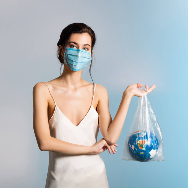 молодая женщина в медицинской маске держит пластиковый пакет с глобусом на синий, экологическая концепция  - Фото, изображение