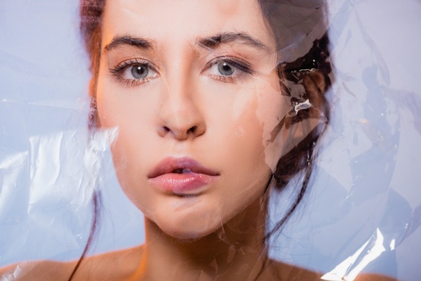 brunette young woman looking at camera through polyethylene on grey, ecology concept  - Fotografie, Obrázek