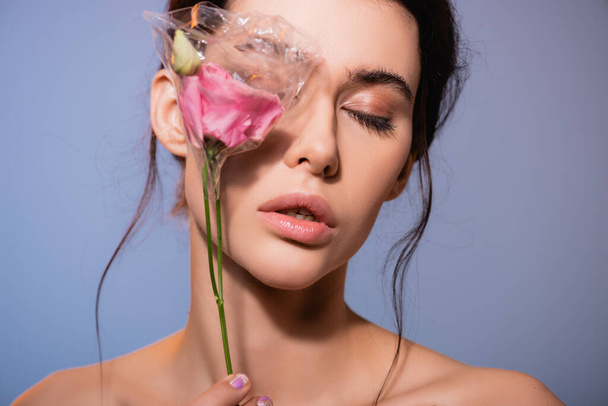 young and naked woman covering eye while holding flowers in polyethylene isolated on grey, ecology concept  - Photo, Image