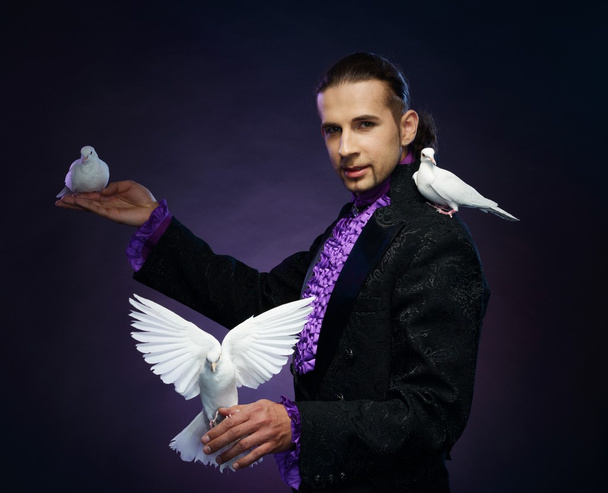 彼の訓練を受けた白い鳩のステージ衣装で若いハンサムなブルネット魔術師男 - 写真・画像
