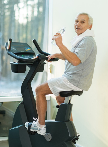 Hombre mayor con botella de agua en una bicicleta en un gimnasio
 - Foto, Imagen