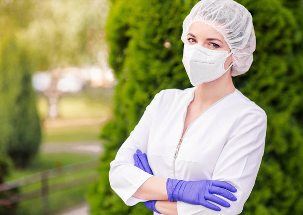 Een mooi meisje in een medisch pak, een beschermend masker op haar gezicht, handschoenen en een medische pet vormt zich tegen een achtergrond van groen. Het meisje staat met gekruiste armen en kijkt in de camera. Horizontale foto - Foto, afbeelding
