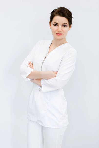 Una joven hermosa y linda con un peinado de moño, un traje médico y brazos cruzados posando contra una pared blanca. Foto vertical - Foto, Imagen