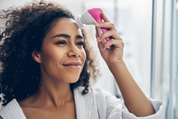 Dreamy woman applying an electric facial cleansing brush - Photo, Image