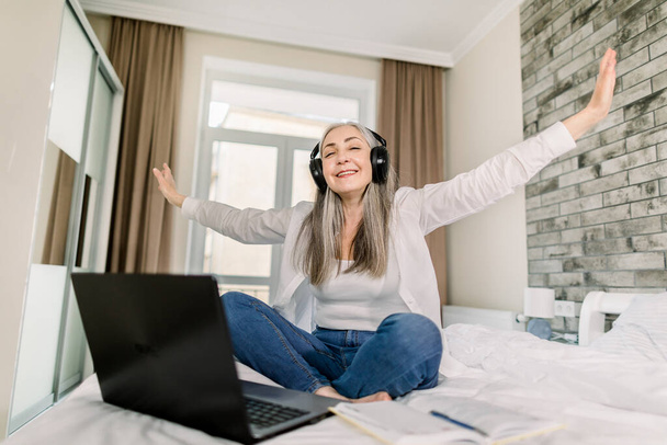 Horizontal indoors home shot of joyful grey haired senior woman wearing headphones and enjoying listening to the music, sitting on the bed with arms outstretched - Photo, Image