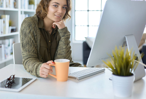 Attractive smiling woman sitting at office desk, holding a cup of coffee, she is relaxing and looking away - Foto, Imagen