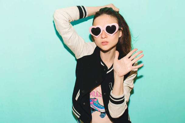 young hipster beautiful woman, modern swag style, jacket, funny heart sunglasses, against blue wall, non-isolated, denim jeans, fashion trend summer, cool face expression - Photo, image