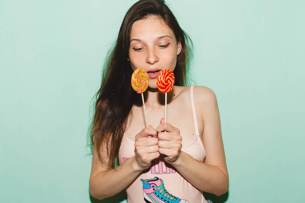 young beautiful hipster woman posing against blue wall, denim vintage style, pink swimsuit, blue jeans, holding eating lolly pop candy sexy - Foto, imagen