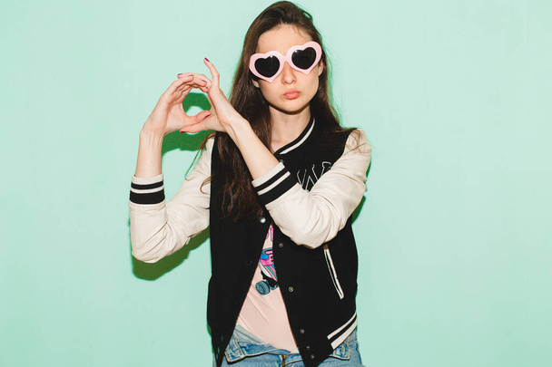 young hipster beautiful woman, modern swag style, jacket, funny heart sunglasses, against blue wall, denim jeans, fashion trend summer, cool face expression - Photo, image