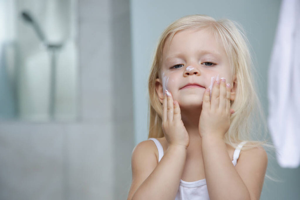 Adorable blonde caucasian toddler girl applying cream on her face - Foto, imagen