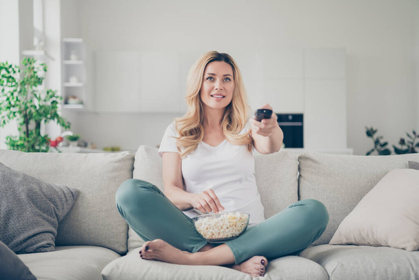 Photo of pretty domestic beautiful lady sit couch eat popcorn remote control change channel watch tv weekend quarantine stay home good mood legs crossed living room indoors - Fotoğraf, Görsel