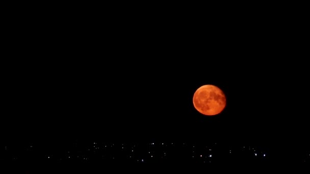 Heldere rode oranje maan oprijzend op donkere nachtelijke hemel boven stadslichten. Mystieke nachtelijke zwarte hemel met grote maan, donkere rust - Video