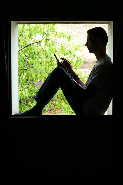技術を使ってる人。窓の中に男のシルエット,彼の手の中にスマートフォンと.背景は意図的に太陽の下で植生とぼやけている. - 写真・画像