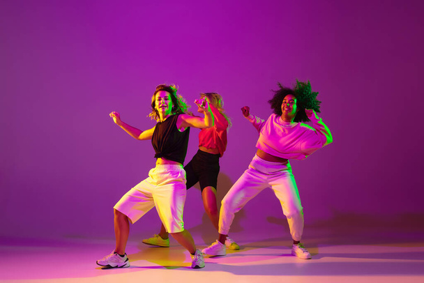 Sportive girls dancing hip-hop in stylish clothes on gradient background at dance hall in neon light - Фото, изображение