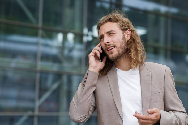 Young businessman talking on the phone, standing on city street near business center. Successful manager makes a call. Portrait of handsome curly smiling man in casual wear holding smartphone - Foto, imagen