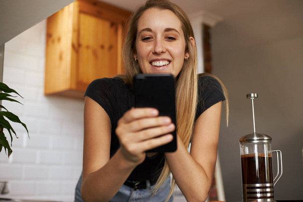 Молодая женщина с помощью смартфона опираясь на кухонный стол делает кофе - Фото, изображение
