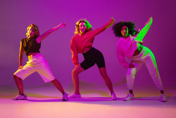 Sportive girls dancing hip-hop in stylish clothes on gradient background at dance hall in neon light - Zdjęcie, obraz