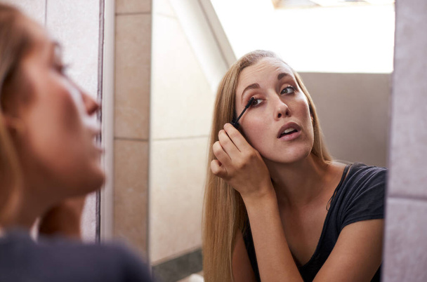 Young blonde woman applying makeup in mirror in bathroom - Фото, изображение