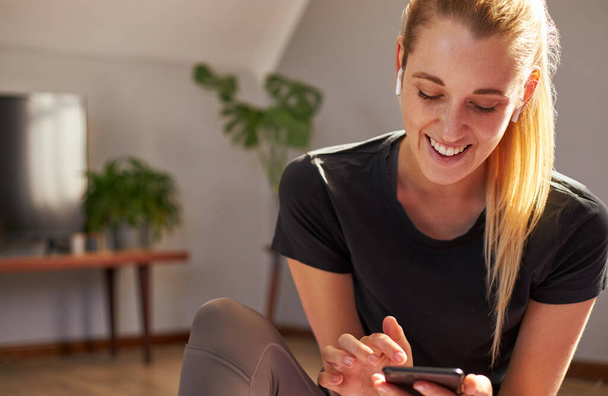 Mujer joven usando el teléfono inteligente mientras hace ejercicio en casa con auriculares en. - Foto, imagen