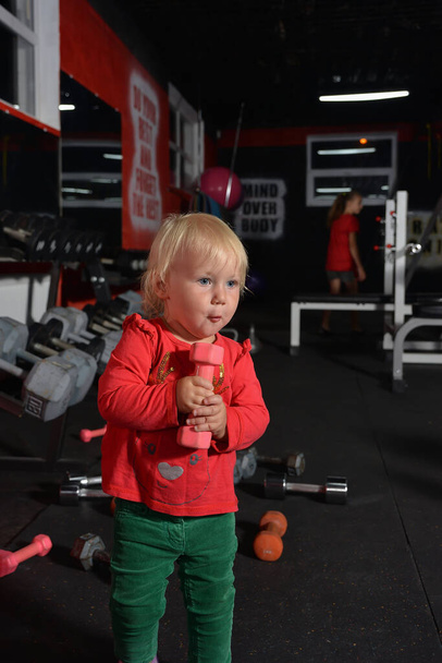 Un petit enfant tient un haltère dans ses mains. Petite fille va faire du sport dans la salle de gym. - Photo, image