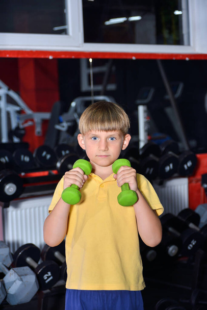 Le garçon d'âge préscolaire tient des haltères dans ses mains. L'enfant va faire du sport dans la salle de gym. - Photo, image