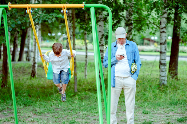 dad in the playground. Dad looks at the phone. The boy is riding on a swing. - Foto, imagen