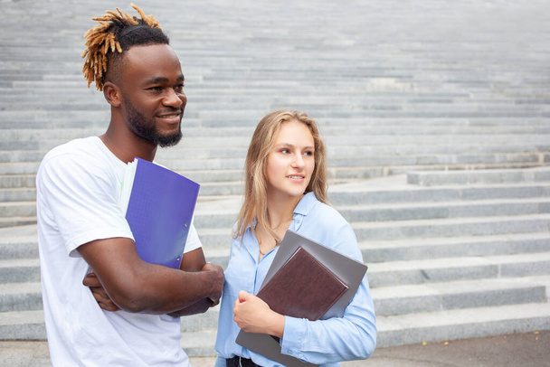 Educação e conceito de pessoas. Retrato de dois estudantes multiétnicos sorridentes, posando ao ar livre. Afro-americanos do sexo masculino e europeu do sexo feminino na cidade, estudar para seus exames on-line, olhando para longe. - Foto, Imagem