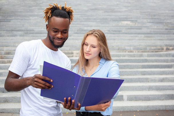 Concepto de educación y personas. Retrato de dos estudiantes multiétnicos, posando al aire libre. chico afroamericano y chica europea en el parque, mirando el cuaderno y estudiar para sus exámenes en línea. - Foto, Imagen