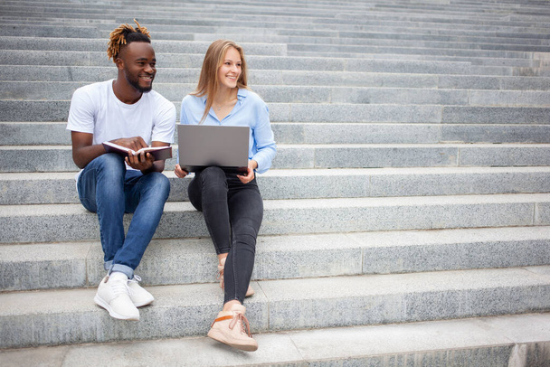 Bildungs- und Personenkonzept - junge europäische Frauen und afroamerikanische Männer. Studenten sitzen auf Treppen im Park, lächeln und bereiten sich auf Online-Prüfungen vor, schauen weg. - Foto, Bild