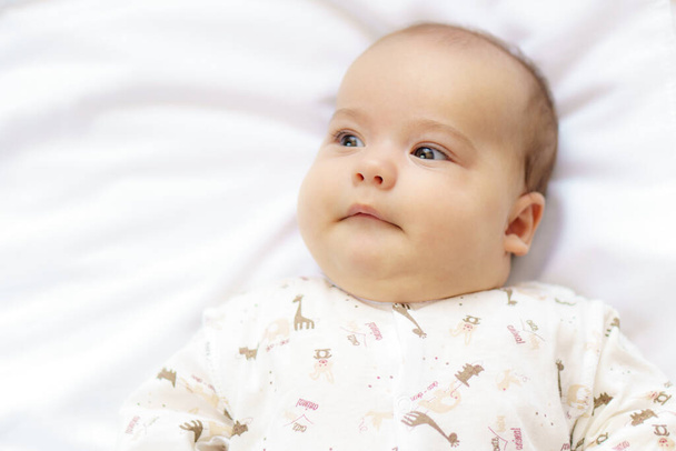 Little newborn baby in a white bodysuit lying on the bed. Top view of a newborn baby smiles on a white warm blanket. The newborn is awake looking around indoors - Photo, Image