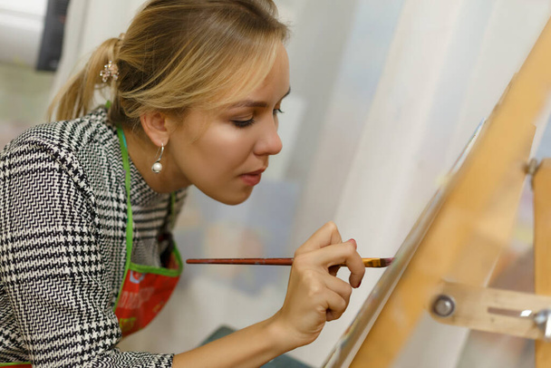 A beautiful blonde girl artist paints a picture on a canvas with oil paints. - Foto, Imagen