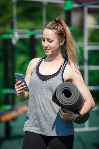 Fröhliche junge Frau hat Trainingspause, posiert mit Fitnessmatte, hält Smartphone, trägt Sportbekleidung. Konzept der guten körperlichen Form und gesunden Lebensweise. - Foto, Bild
