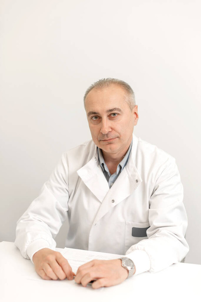 Portrait of a professional male doctor in a white coat sitting at a table. - Fotó, kép