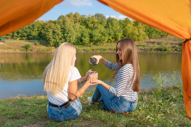 Camping on lake shore at sunset, view from inside camping tent. Two beautiful girls enjoy nature and drink hot tea in front of tent. - Фото, изображение