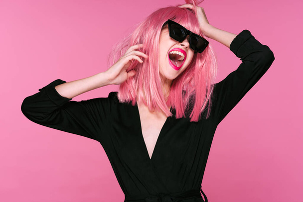Grabado en estudio. Joven chica feliz en peluca rosa y gafas de sol sobre fondo rosa - Foto, imagen