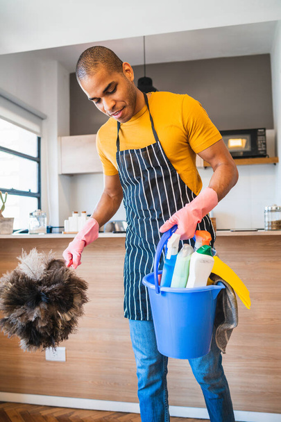 Portrait of young latin man holding a bucket with cleaning items at home. Housekeeping and cleaning concept. - Valokuva, kuva