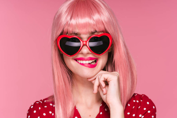 hermosa mujer en peluca rosa y gafas de sol en forma de corazón sobre fondo rosa  - Foto, Imagen
