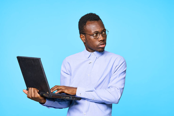 Νεαρός άνδρας με γυαλιά με φορητό υπολογιστή κοιτάζει μακριά απομονωμένος σε μπλε φόντο  - Φωτογραφία, εικόνα