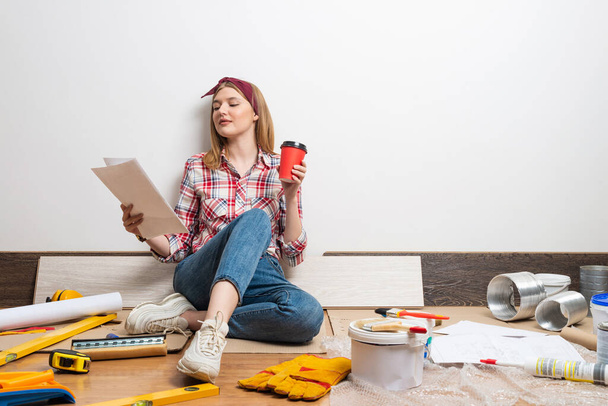 Happy pretty girl sitting on floor with paper blueprint. Home remodeling and house interior redesign. Construction tools and materials for building. Young woman studying renovation project of flat. - Fotoğraf, Görsel