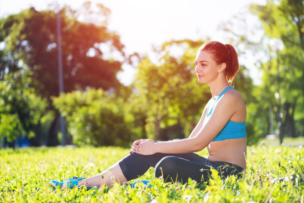 Beautiful smiling girl in sportswear relax in park. Young woman with sitting on green grass after training. Meditation outdoor at sunny summer day. Morning exercises and healthy lifestyle. - Foto, Imagen