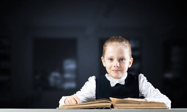 Smiling little girl sitting at desk with open book. Research and education in school. Happy schoolgirl studying in library. Clever girl in schoolwear reading book on dark blurred background. - Foto, imagen