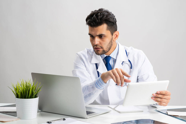 Mladý arabský internista pracující s tabletovým počítačem. Lékař v bílých lékařských šatech se stetoskopem sedí za stolem s laptopem. Odborná lékařská konzultace a léčba na moderní klinice. - Fotografie, Obrázek