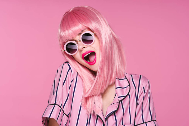 Młoda piękna kobieta w różowej peruce i okularach przeciwsłonecznych z otwartymi ustami na różowym tle - Zdjęcie, obraz