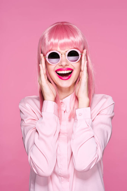 Jonge mooie vrouw in roze pruik en zonnebril glimlachen op roze achtergrond - Foto, afbeelding