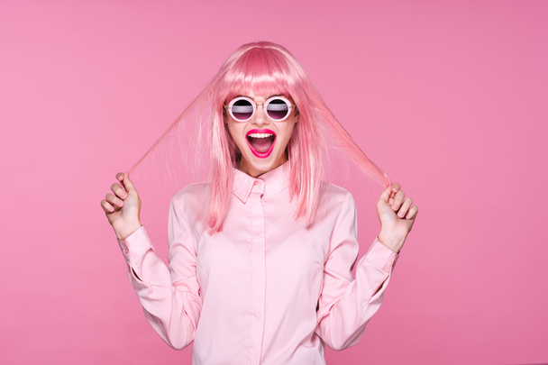 Νεαρή όμορφη γυναίκα σε ροζ περούκα και γυαλιά ηλίου είναι ευτυχισμένη σε ροζ φόντο - Φωτογραφία, εικόνα