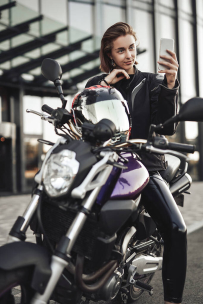 Egy motoros dzsekis lány ül egy lila motoron, és nézi a telefonját. - Fotó, kép
