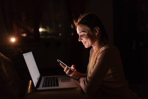 woman working remotely on her laptop in the evening during the quarantine to avoid the spread of coronavirus. Holding smartphone - Foto, afbeelding
