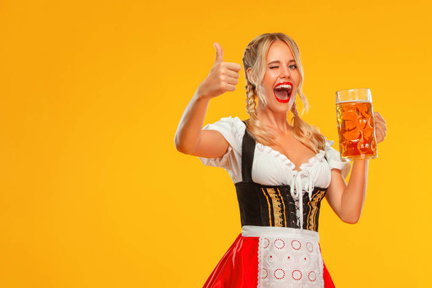 Joven chica sexy Oktoberfest camarera, con un dirndl tradicional bávaro o alemán. Mujer sirviendo tazas de cerveza grandes con bebida aislada sobre fondo amarillo. - Foto, Imagen